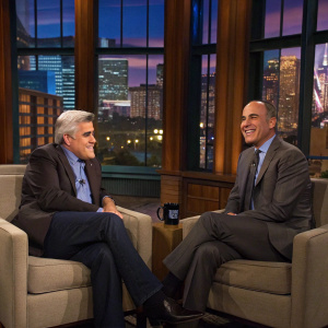 Jay Leno and Matt Lauer in a cozy studio. Large windows showcasing a bustling cityscape. Warm lighting, plush chairs. Friendly and relaxed demeanor.