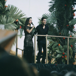 Lana Parrilla and Paul Wesley on the vibrant Teen Choice Awards stage. Lana in a stunning black lace dress with sparkling embellishments. Paul in a sleek black shirt. The stage adorned with vibrant tropical imagery.