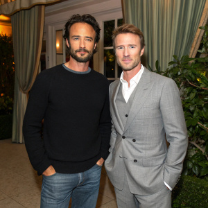 Rodrigo Santoro and Nick Styne at the annual Film Awards. Rodrigo, dressed in a black sweater and jeans, takes a casual pose. Nick, impeccably groomed in a gray suit, stands formally. Background is blurred greenery and curtain.