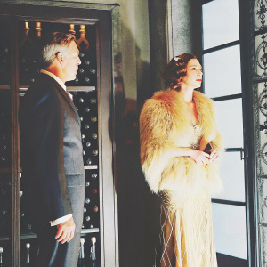 Carlo Ancelotti and Mariann Barrena at a luxurious gala. Carlo in dark suit, Mariann in a lavish dress with golden hair and fur-like details. Elegant wine-themed backdrop. Softly lit room.