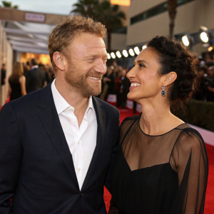 Kevin McKidd and Indira Varma at a gala event. Kevin in a dark suit and white shirt, beard. Indira in a black dress with sheer overlay. Warm smiles, candid moment on red carpet.