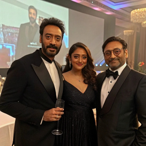Ajay Devgn and Abhishek Pathak, both in formal wear, stand beside Ishita Dutta who is holding a glass. They are at a glamorous awards ceremony with a large screen backdrop.