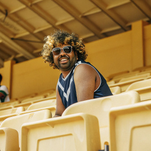 Lasith Malinga, Sri Lankan cricket player, posing for a candid photo at the stadium. He sports multicolored curly hair and shades. Navy blue sleeveless jersey with white accents and brand logos. Background is the vibrant crowd and seating area.