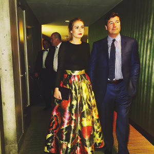 Kyle Chandler and his daughter Katherine at a gala. She's in a vibrant printed skirt and black top. He wears a navy suit and striped shirt. Formal setting, dim lighting.