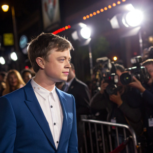 Dane DeHaan at a Hollywood premiere. Dapper in a blue suit and white shirt. Bright lights and paparazzi in the background.