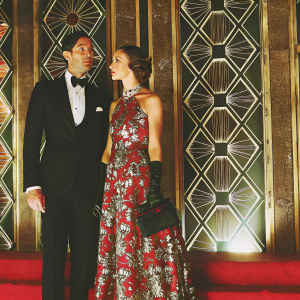 Adrien Brody and Lara Lieto attending the Oscars after-party. He's in a black tuxedo, she's in a sparkling floral gown. Patterned background with gold accents.