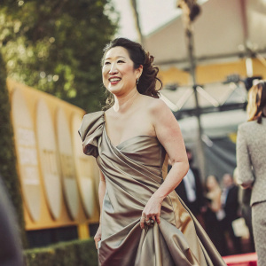 Sandra Oh, with a radiant smile, dressed in a brown silk gown at a glamorous event. The backdrop is a blend of gold and yellow hues, with numerous logos of sponsors and organizers. The setting is a high-profile awards ceremony.
