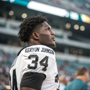 Kerryon Johnson, wearing white and black jersey with number 34, frowning. Dark, messy hair. Stadium background, crowd blurred.