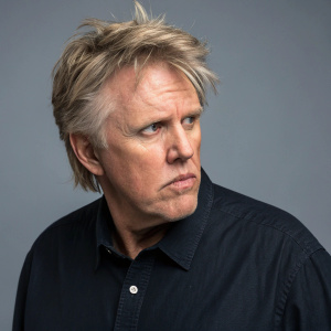 Close-up of Gary Busey, Caucasian actor, in a tense, focused pose. Dark button-up shirt. Solid gray background.
