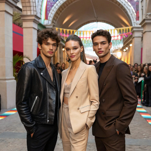 Desire Cordero in a black leather jacket, Josephine Skriver in a beige blazer, Xavier Serrano in a dark brown suit. They are at a trendy outdoor event with an archway and crowd visible in the background.
