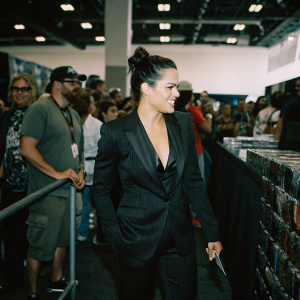 Michelle Rodriguez at Comic-Con. Wearing a stylish black suit, dark hair up in a bun, radiant smile. Crowded convention center backdrop.