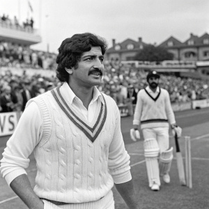 Pakistani cricketer Shafqat Rana, with his mustache, in the middle of a bustling cricket stadium. He's dressed in a white V-neck sweater, posing with a teammate. The picture is in black and white.