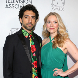 Ricky Paul Goldin and Beth Ehlers at the Television Critics Association Press Tour. Ricky, a latino actor, dressed in a black suit with a vibrant floral tie. Beth, a blonde actress, in a flowing emerald green dress. Both stand beside a white backdrop with event signage.