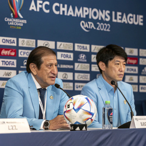 Press conference for AFC Champions League 2022 in Doha, Qatar. Ramon Diaz and Sang Hyun-Soo sit at a table with soccer-themed backdrop. The table is adorned with microphones and a soccer ball centerpiece. Both men wear light blue suits.