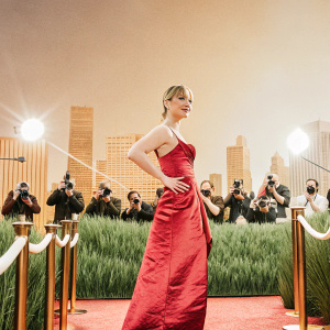 Saoirse Ronan at a film festival, wearing a red velvet gown, posing gracefully. Crowded event, flashing cameras, city skyline backdrop