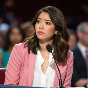 Lulu Antariksa seated at an awards ceremony. Serious expression. Asian ethnicity. Medium-length brown hair. Soft natural makeup. Wearing a white blouse and pink blazer. Standard microphone in front of her. Blurred indoor setting with audience visible.