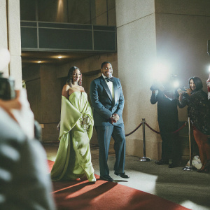 Teasha Bivins and Michael Bivins at a glamorous Hollywood Awards Show. Teasha in a flowing emerald green gown, Michael in a navy blue tuxedo. Background filled with flashing cameras and paparazzi.