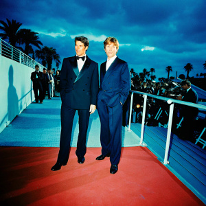 Tom Cruise and Robert Redford at the Cannes Film Festival premiere of Lions for Lambs. Both dressed in elegant formal wear. Tom in a classic black tuxedo. Robert in a sophisticated navy suit. Against a bustling red carpet lined with fans and photographers.