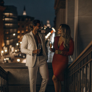 Mariano di Vaio and Eleonora Brunacci at a high-profile fashion event. Mariano in a white suit, Eleonora in a vibrant red dress. The scene is set against the backdrop of a bustling city skyline, with flashing neon lights.