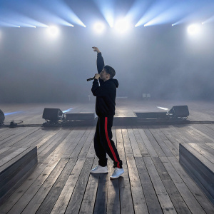 Rich Brian, male performer, intense stage presence. Wears black hoodie, black pants with red stripe, white sneakers. Bright blue stage lights. Large indoor venue, wooden floor, with stage equipment visible in background.