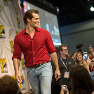 Henry Cavill attending a Comic-Con event. Wearing a red shirt and jeans. Surrounded by excited fans and convention attendees.