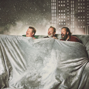 James Corden and guests Bradley Whitford, Brett Gelman laughing heartily. Behind the scenes of the Late Late Show studio, filled with mock cityscape backdrop and gray couch setup.