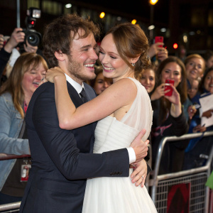 Sam Claflin and Laura Haddock, both white, hugging at premiere party. Fans cheering and taking pictures in the background