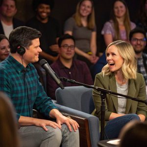 Mike Schur and Kristen Bell engaged in an animated discussion during a podcast recording session. A bustling audience watches intently in the background.