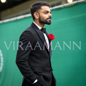 Virat Kohli, cricket star, standing in front of a blurred green screen. Wearing a formal black suit and red rose in lapel. Text overlay reads 'Viraamann'