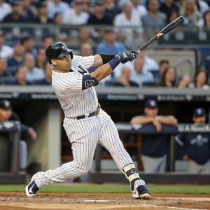 Gary Sanchez, wearing New York Yankees jersey, mid-swing at baseball game. Crowd and stadium blurred in background.
