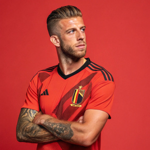 Toby Alderweireld, Belgian footballer, styled haircut, stubble, posing in an adidas jersey with Belgium's emblem. Solid red background.