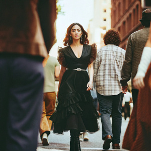 Shay Mitchell, Asian actress, walking down a busy city street. Wearing a black dress with frilly details and a matching belt, elegant black boots. She has a serious expression.