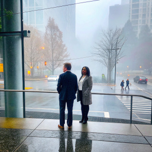 Patrick Duffy and Carlyn Rosser, both Caucasian, standing outside a modern office building. Patrick in a dark suit, Carlyn in a white blouse with a checkered blazer. A busy city street backdrop.
