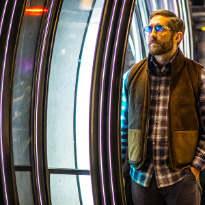 Jake Gyllenhaal in a professional photo shoot. Wearing a brown vest over a plaid shirt, blue tinted glasses, and a neatly trimmed beard. The background is a simple, smooth white