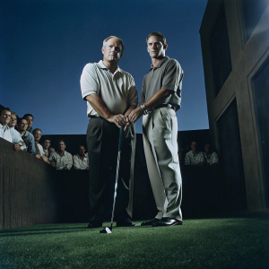 Jack Nicklaus and Drew Brees standing on a sunny green golf course. Spectators in the background watch intently.