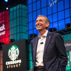 Howard Schultz, ex-Starbucks CEO, smiling for camera at a tech conference. Dynamic backdrop with LED screens and brand logos.