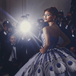 Zendaya at the Met Gala. Wearing an extravagant blue dress with peacock feathers. Crowded backdrop with flashing cameras.