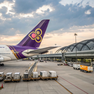A luxurious Airbus A380, painted in Thai Airways livery, parked at the bustling Suvarnabhumi International Airport. The aircraft's tail features a vibrant purple and gold emblem. Surrounding airport facilities, including jetways and cargo containers, are visible in the background.
