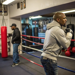 Floyd Mayweather during a boxing training session. Sweatshirt, jeans, standing beside a boxing ring. Trainer in the background. Urban boxing gym