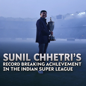 Sunil Chhetri, in celebration of his milestone goal, poses with the trophy. The blurred background features a packed stadium and green soccer field. The text reads 'Sunil Chhetri's record-breaking achievement in the Indian Super League.'