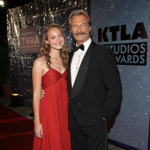 Perry King and his charming daughter Louise, posing for the cameras. Perry, sporting a classic mustache, stands next to Louise, elegantly dressed in a long, red evening gown. The backdrop is the glittering KTLA Studios in downtown Los Angeles during the annual Stuntman Awards ceremony.