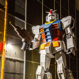 Gundam RX-78-2, standing in an action pose, set against a black backdrop. The mecha is predominantly grey with red and blue accents, detailing its armor and weaponry. Its shadow stretches across the dark background.