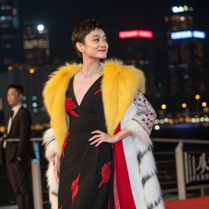 Sun Li, Chinese actress, elegant in a bold black dress with red accents and a yellow and white fur stole. Standing confidently at a gala event with a blurred cityscape in the background.