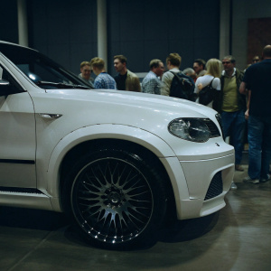 At a bustling car show, the Hamann BWW X5 M50d, a gleaming white chariot, stood out. Its wheels, a marvel of intricate design, shimmered with a dark, metallic finish and a central emblem. Surrounded by an eager crowd, the vehicle sat on a polished, pristine floor, showcasing its sleek and refined contours under the even lighting.