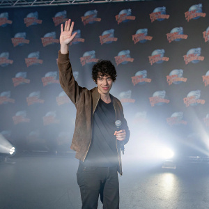 Ian Eastwood, Hispanic actor, with curly dark hair and friendly face. Wearing casual black t-shirt, brown suede jacket. Bright stage lights, colorful backdrop with promotional logos.