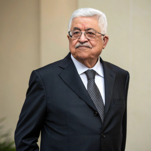 Mahmoud Abbas in a formal gathering, wearing a dark suit and glasses. Neutral colored backdrop