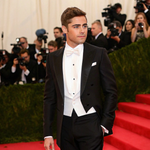 Zac Efron at the Met Gala. Wearing a sharp black tuxedo and white bow tie. Glamorous red carpet, paparazzi in the background.