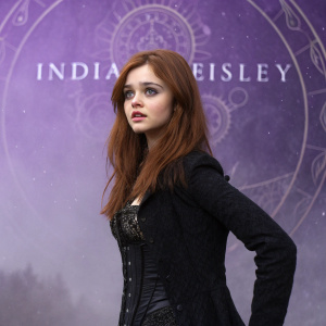 India Eisley, with fair skin and reddish-brown hair, poses somberly. Black outfit with intricate details. Mysterious purple backdrop with ancient text.