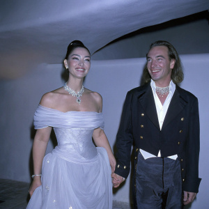 Monica Bellucci and John Galliano at a gala event. Bellucci in elegant white off-shoulder gown with silver necklace. Galliano in stylish black blazer with gold details. Friendly smiles against dimly lit backdrop