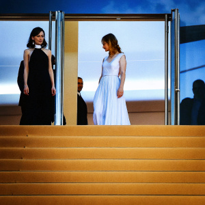 Sibel Kekilli and Nora von Waldstätten attending the Cannes Film Festival. Sibel in dark hair and a black choker, Nora in a flowing white dress. Soft lighting, blurred background.